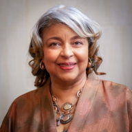 A light brown skinned woman with silver shoulder length hair is smiling wearing a pink blouse. 