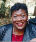A brown skinned woman with short dark hair is smiling. She is wearing a dark blue jacket and a red blouse.